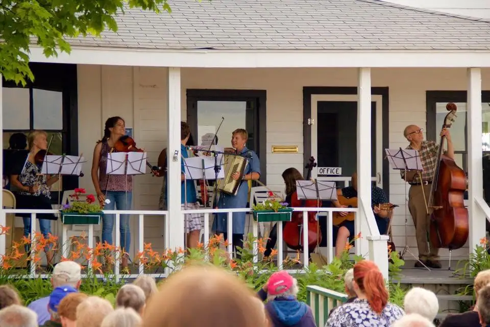 Musicians perform on a porch before a seated audience, with string instruments and an accordion.