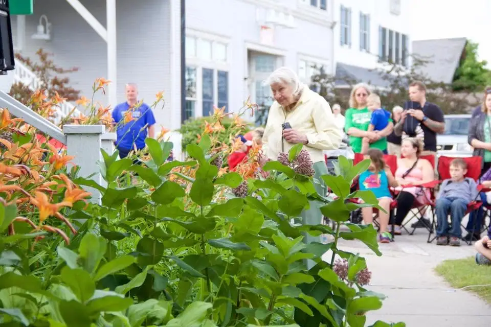 A woman with a microphone stands among flowering plants before an outdoor seated gathering.