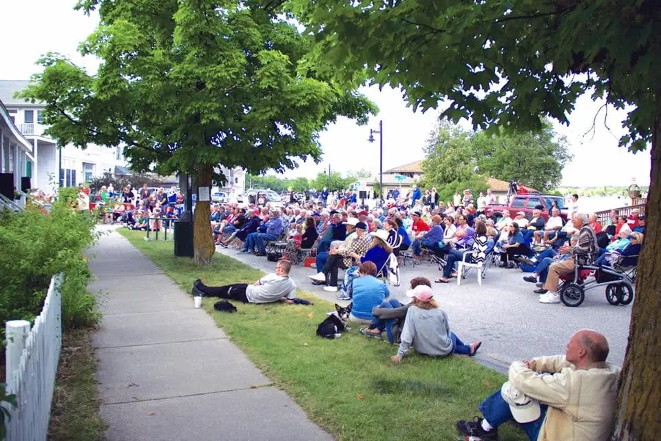 A large seated crowd gathers outdoors beside a sidewalk, lawns, trees, and buildings.