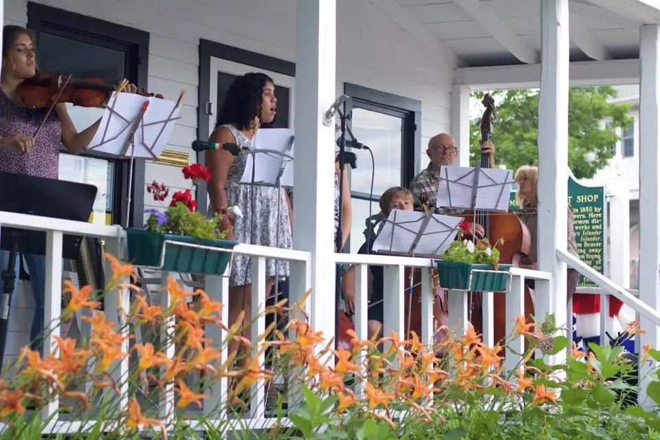 Musicians perform on a flower-decorated porch with violin, upright bass, microphone, and sheet music.