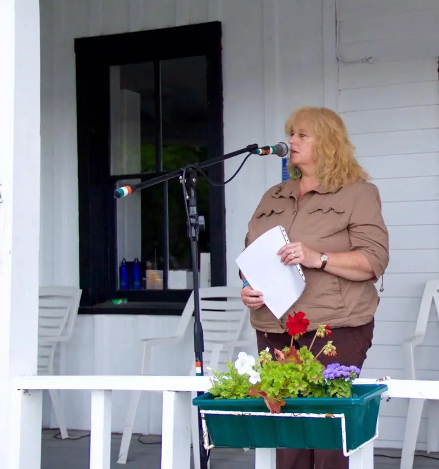 A woman speaks into a microphone while holding papers on a flower-decorated porch.