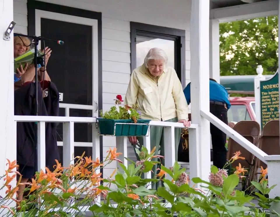 An elderly woman stands on a flower-filled porch beside a microphone and a partially visible historical sign.