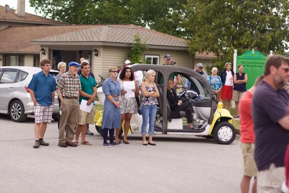 A group gathers beside a golf cart and a green historical marker outside a low building.