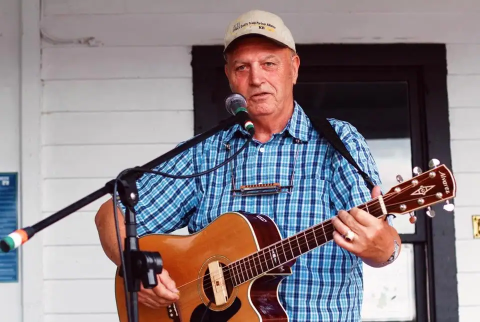 A man sings into a microphone while playing an acoustic guitar, wearing a plaid shirt and baseball cap.