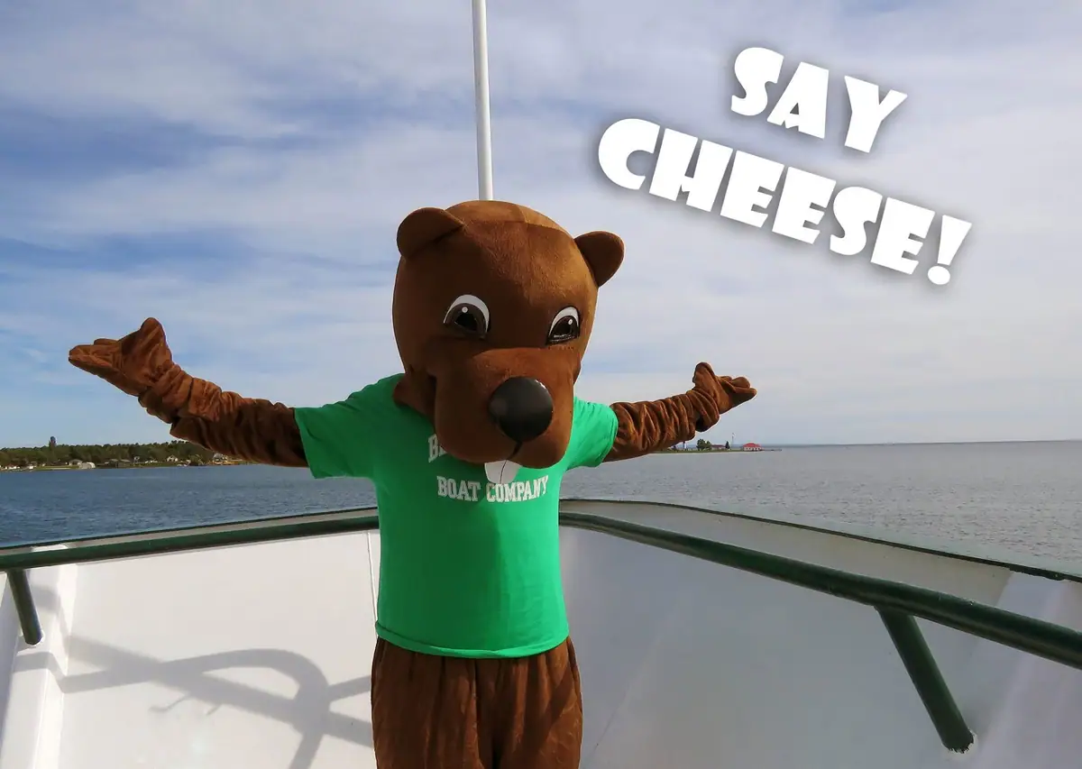 A beaver mascot in a green shirt stands with arms outstretched on a boat deck above the water.