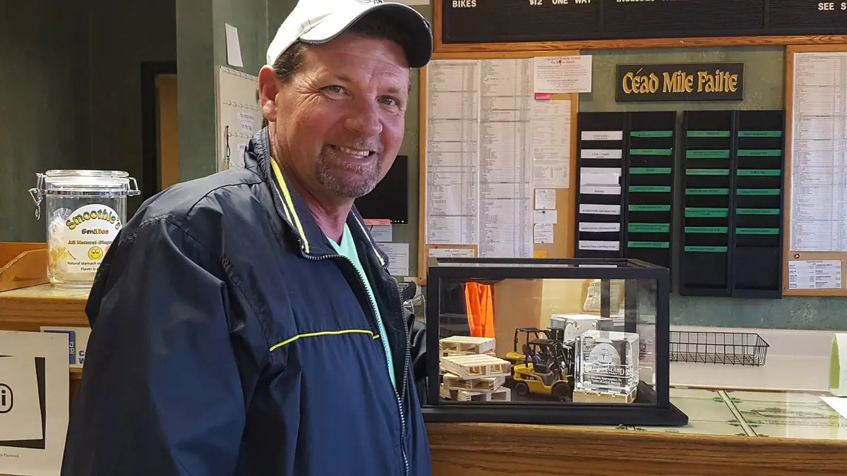 A smiling man in a cap and jacket stands at a service counter beside a bulletin board and display case.