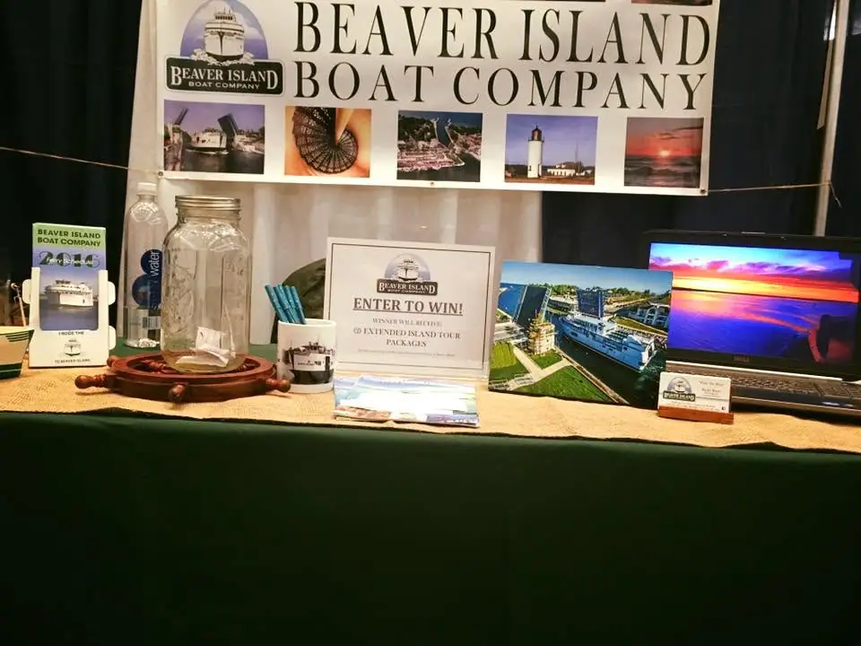 A promotional boat-company booth displays banners, brochures, ferry photographs, a laptop, and a prize sign.
