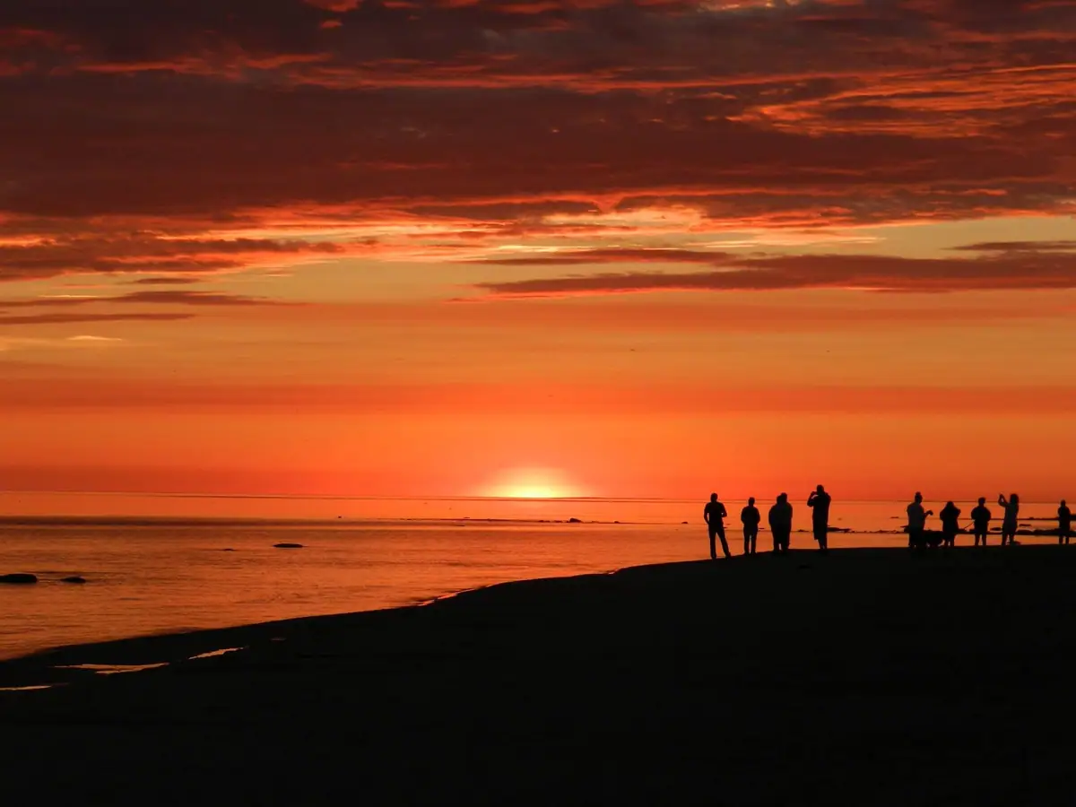 Silhouetted people stand along a shoreline beneath a vivid orange sunset and layered clouds.