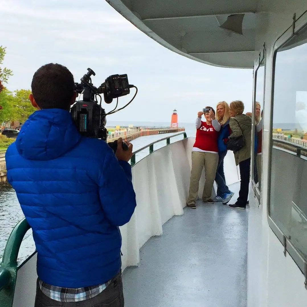 A videographer films three women on a passenger vessel deck near a red lighthouse and pier.