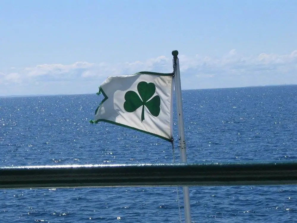 A green-edged white shamrock flag flies from a mast above sunlit open water.