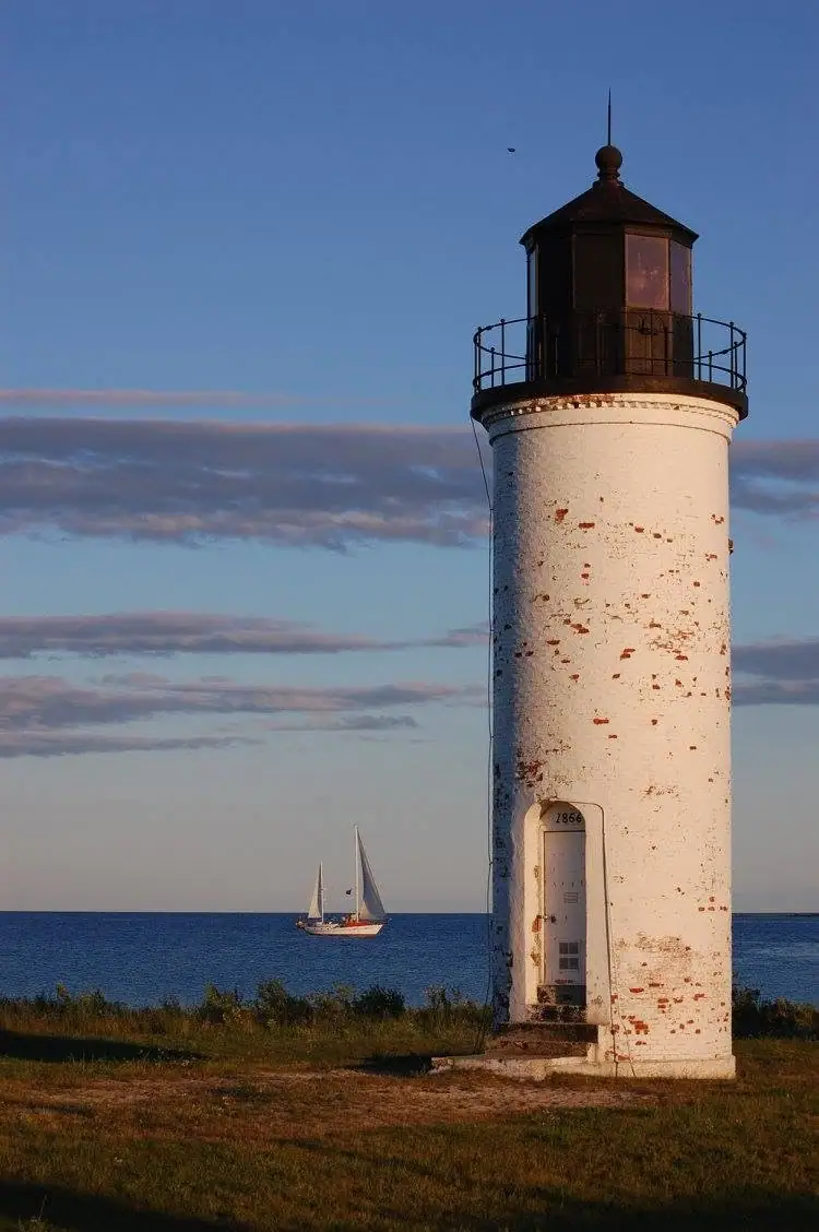 A weathered white lighthouse stands beside the water, with a two-sailed boat visible offshore.