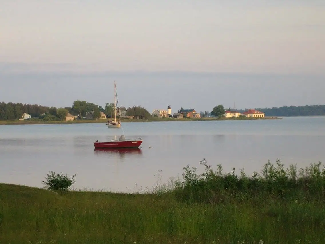 A red boat and a sailboat sit on calm water opposite a tree-lined shore with houses and a tower.