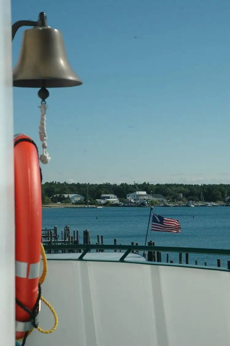 A brass bell and red lifebuoy frame a harbor view with docks, homes, trees, and an American flag.