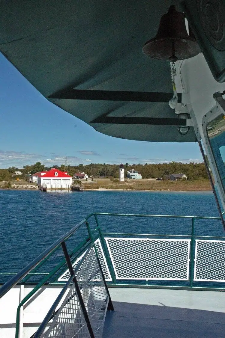 View from a vessel’s deck toward a shoreline with a red-roofed boathouse and a small lighthouse.