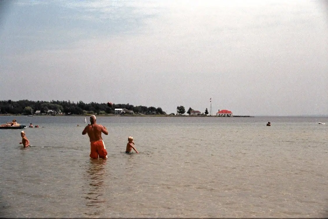 A man and several children wade and play in shallow water while a ball hangs in the air nearby.