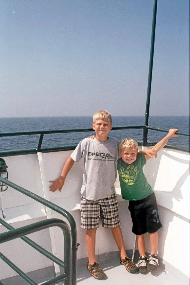 Two boys pose together on a passenger vessel's deck with open water behind them.