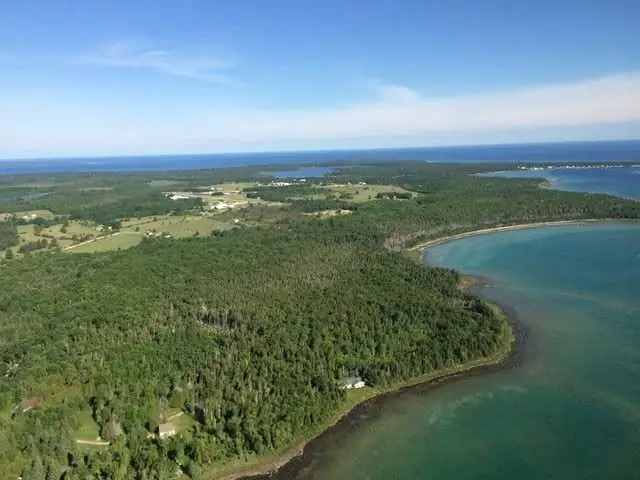 Aerial view of forest, fields, scattered buildings, and a curving shoreline beside blue-green water.