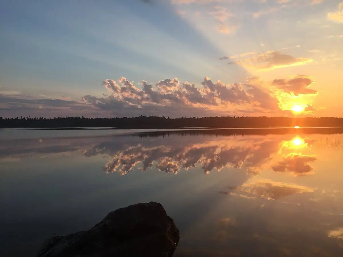Sunset over calm reflective water, with a tree-lined shore and a dark rock in the foreground.