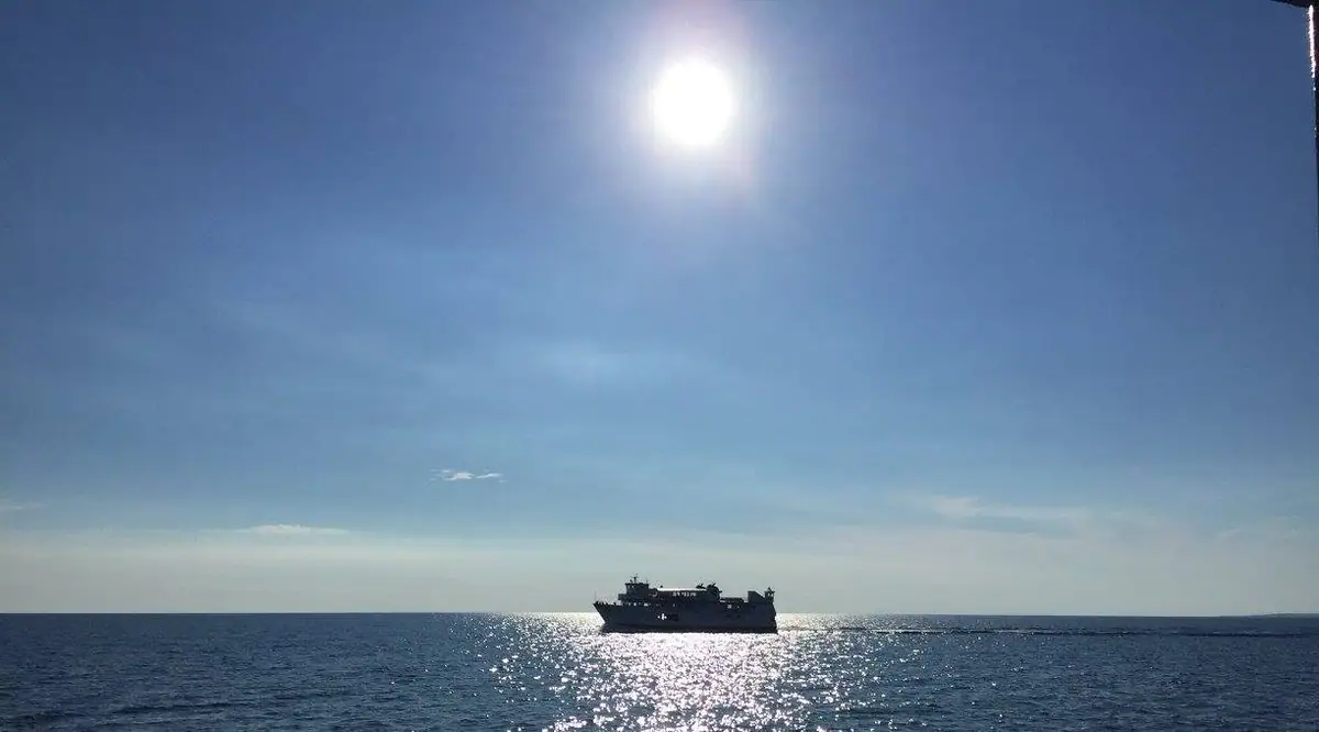 A dark ferry-like vessel crosses open water beneath a bright sun.