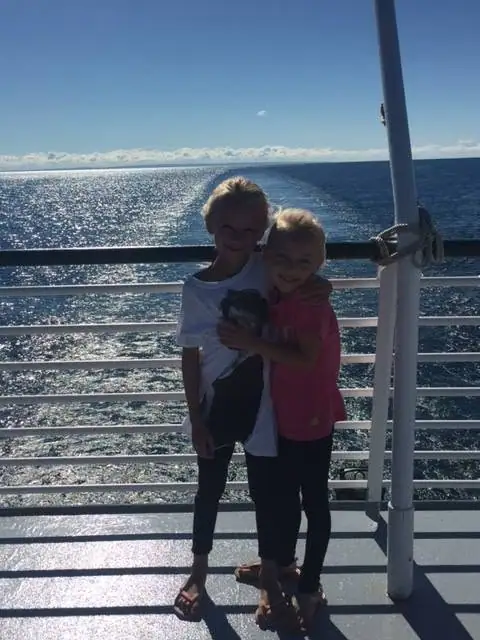Two children hug on a sunlit vessel deck with open water and a wake behind them.