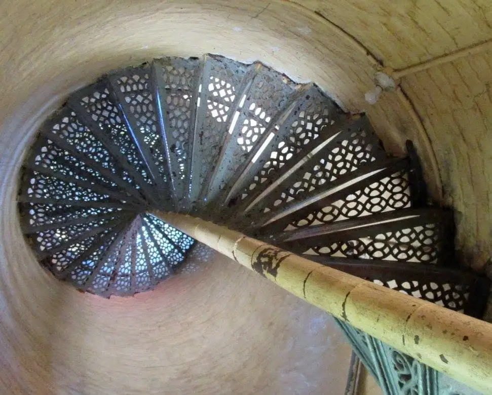 Looking down a spiral staircase with ornate perforated metal steps and a worn bamboo-colored handrail.