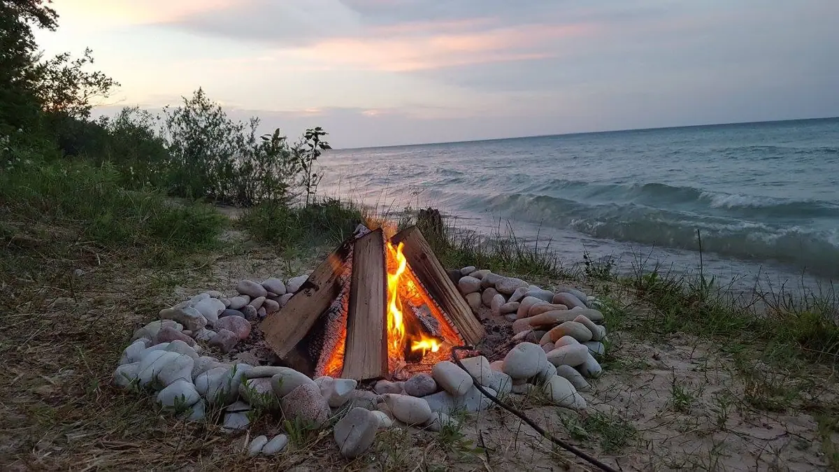 A stone-ringed campfire burns on a sandy shore beside rolling water at dusk.