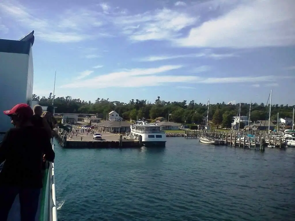 A harbor viewed from a vessel, with people on deck, a white passenger vessel, sailboats, docks, and shoreline buildings.