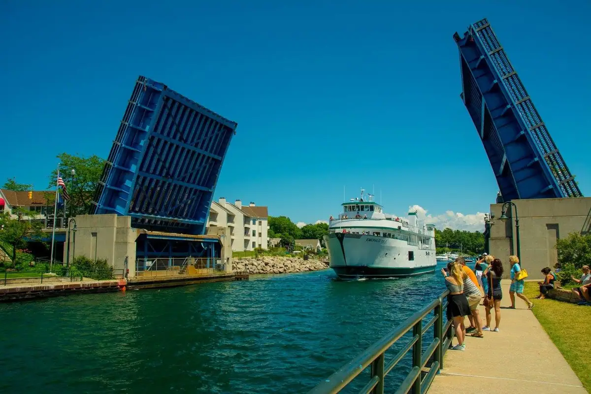 A white ferry passes beneath two raised blue bridge sections while people watch from the waterfront.