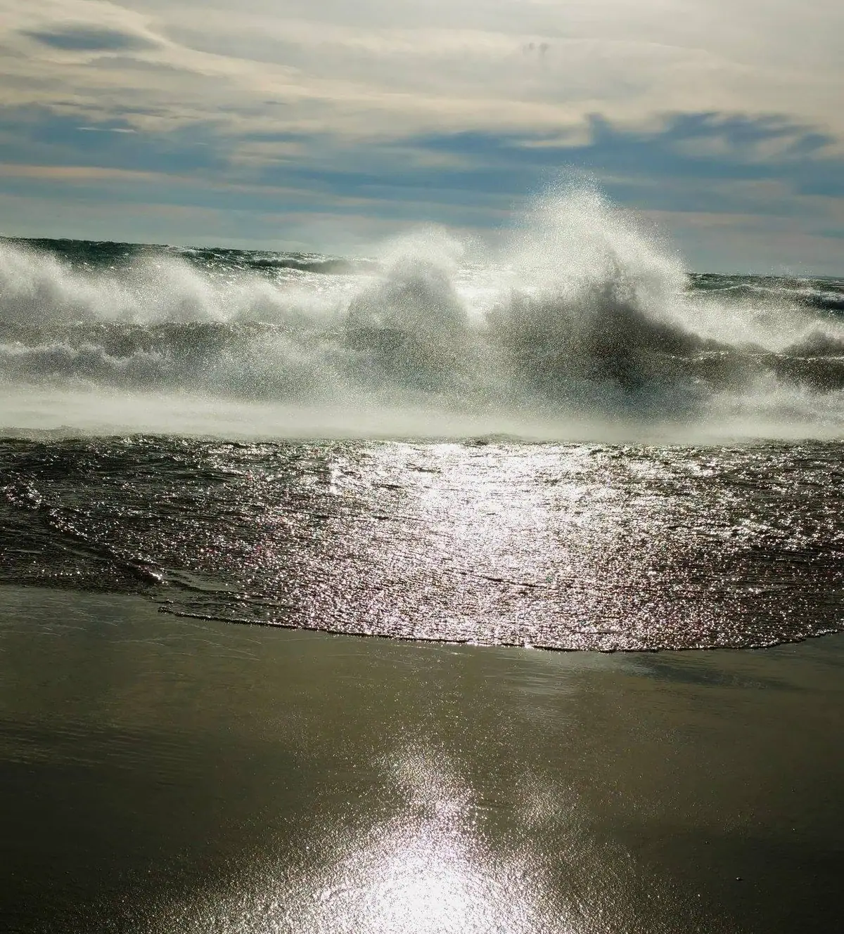 Sunlight glitters across dark, turbulent water as large waves break beneath a cloudy sky.