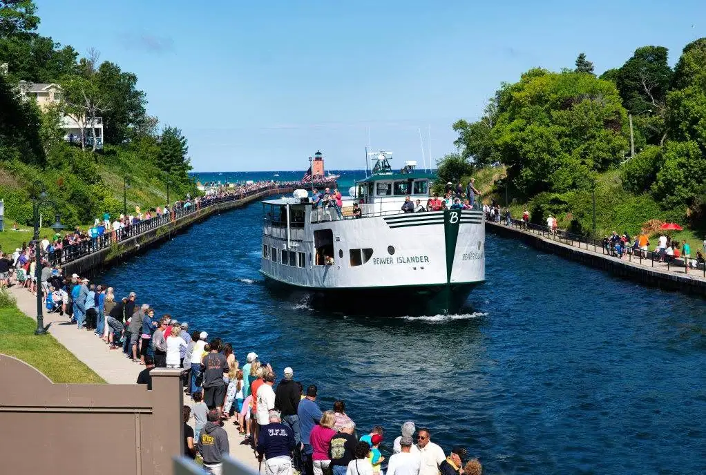 A passenger ferry moves through a narrow channel lined with boardwalks and crowds, with a lighthouse beyond.