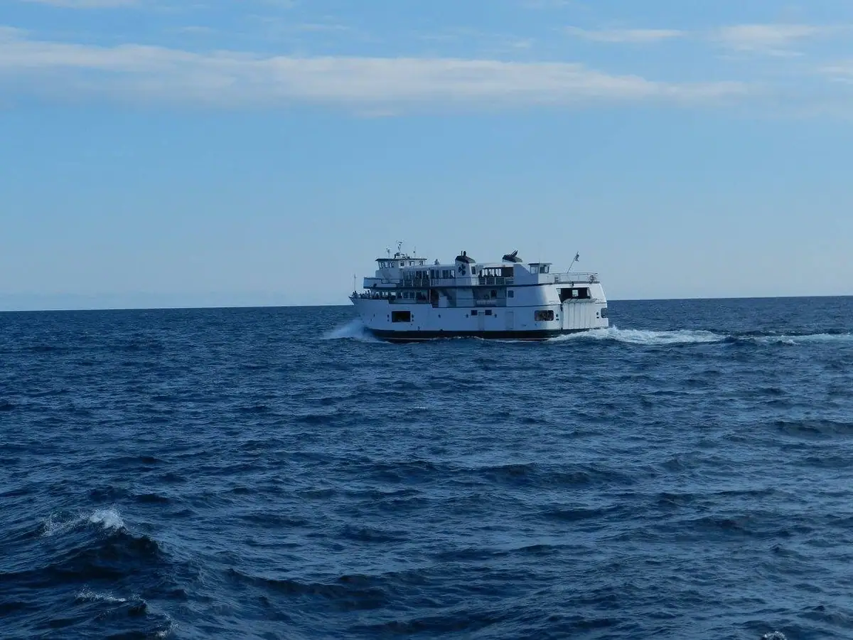 A white multi-deck ferry travels across dark blue water beneath a partly cloudy sky.