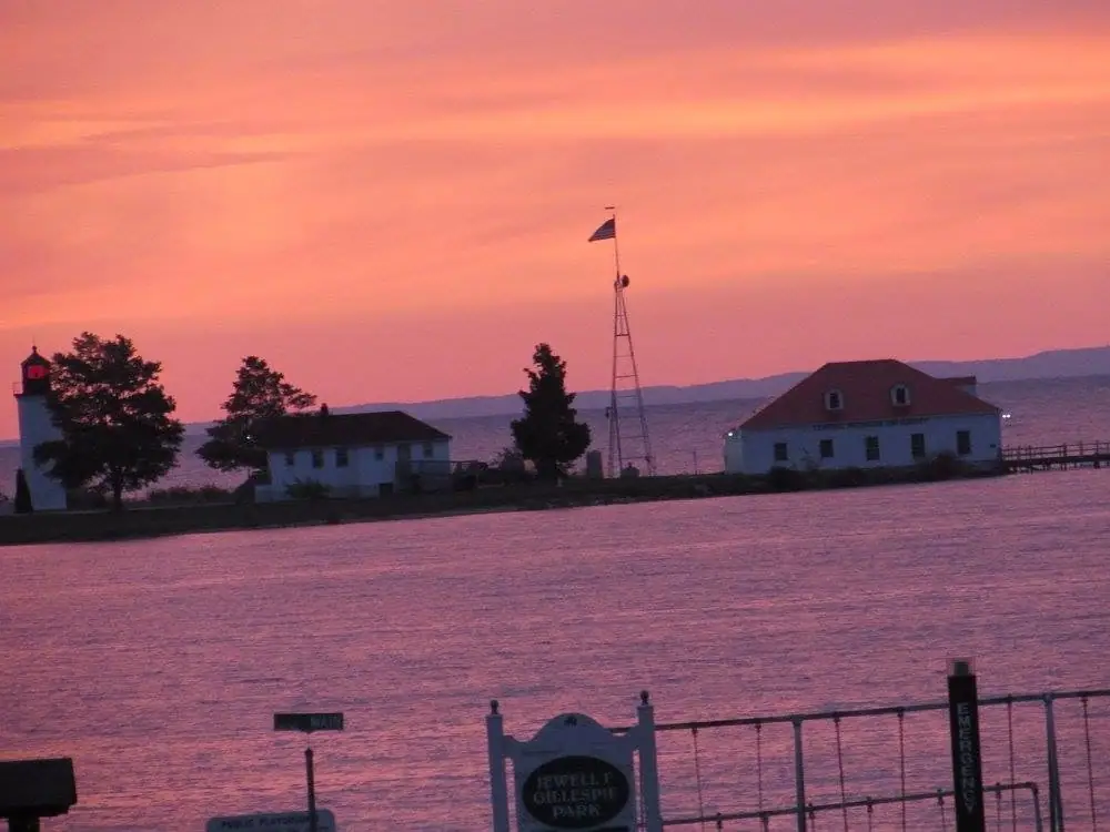 A lighthouse, buildings, flagpole, and pier stand beside calm water under a pink and orange sunset sky.