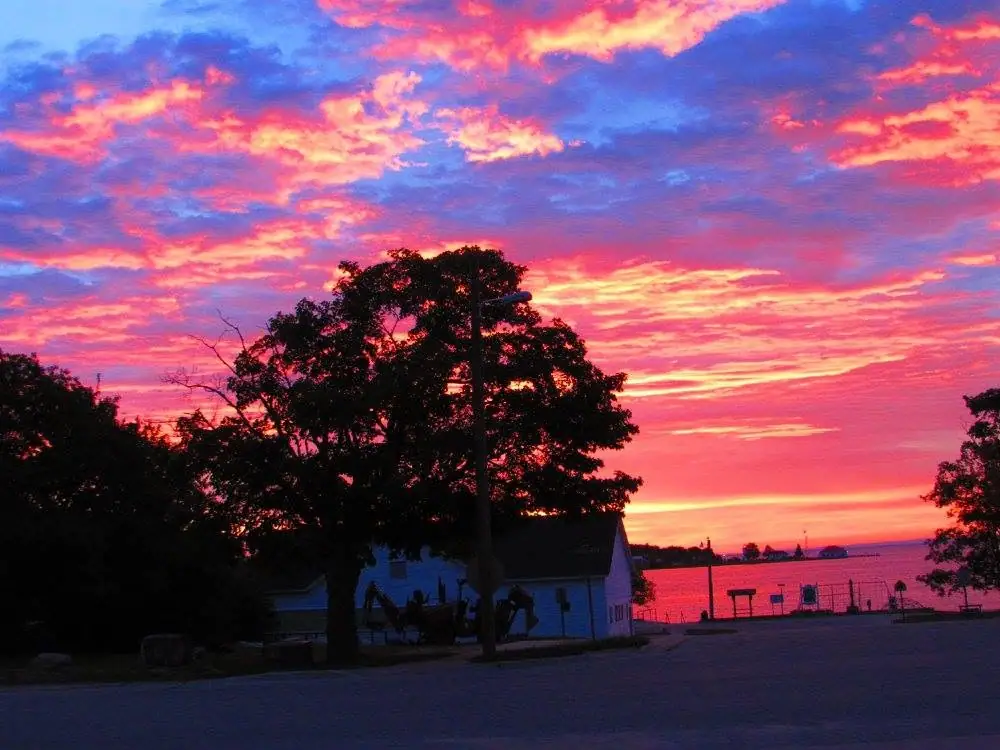 A vivid sunset glows over the water beyond a tree-lined paved waterfront road and small white building.