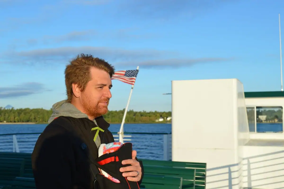 A bearded man holds an infant in a carrier on a boat deck beside an American flag.
