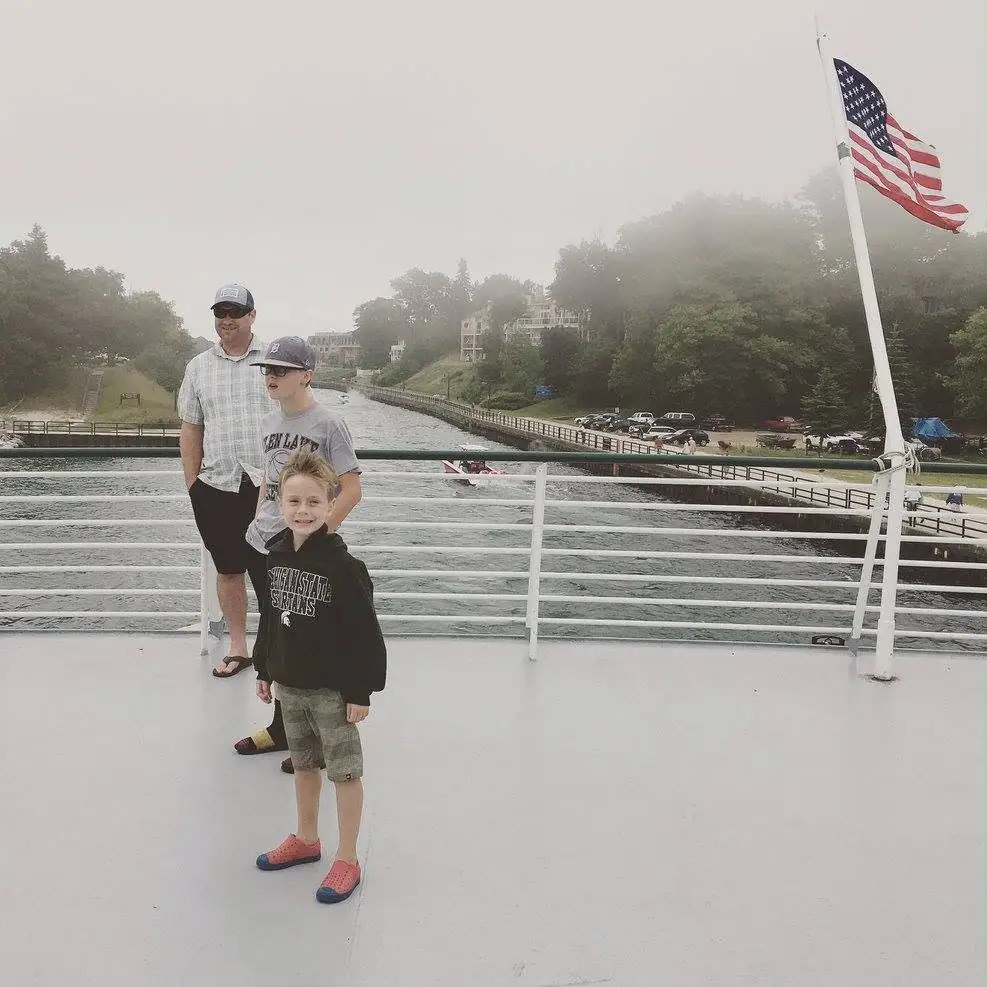An adult and two boys stand on a ferry deck beside an American flag.