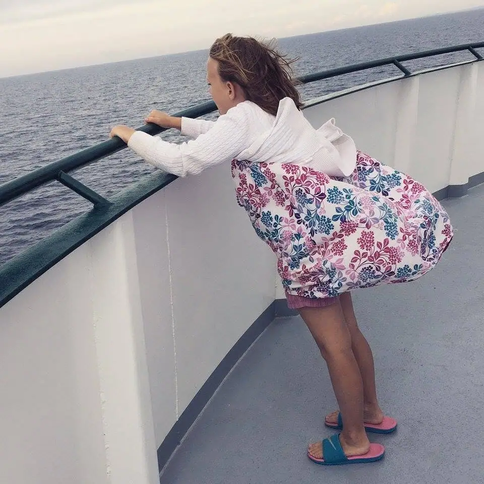 A child in a floral dress leans over a ferry deck railing above open water.