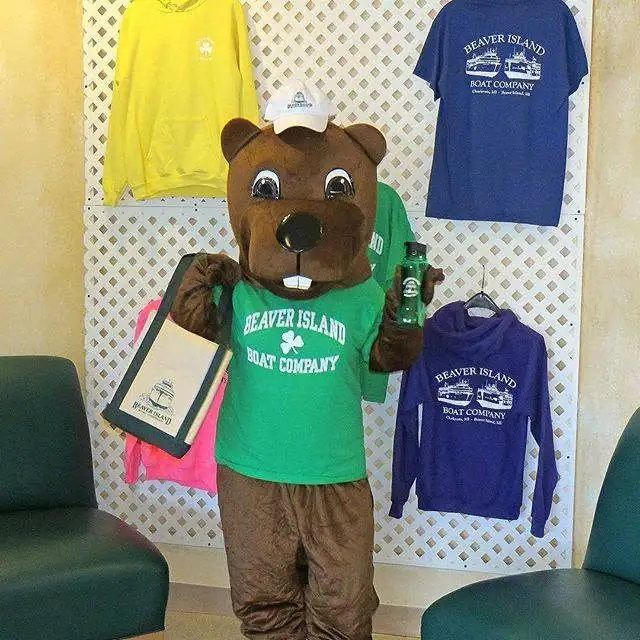 A brown bear mascot in a green shirt and cap stands among displayed apparel, a tote bag, and a bottle.