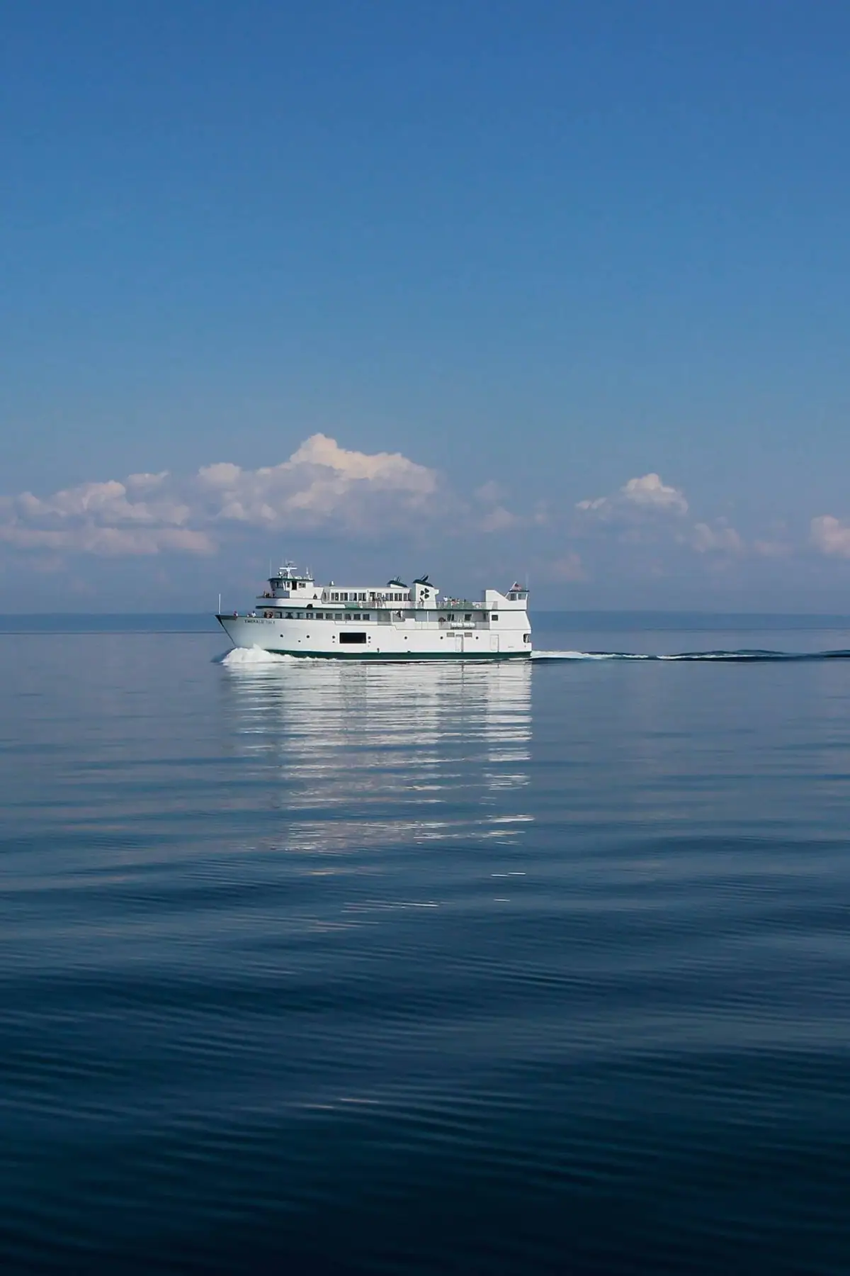A white passenger ferry travels across calm blue water beneath a bright sky with scattered clouds.