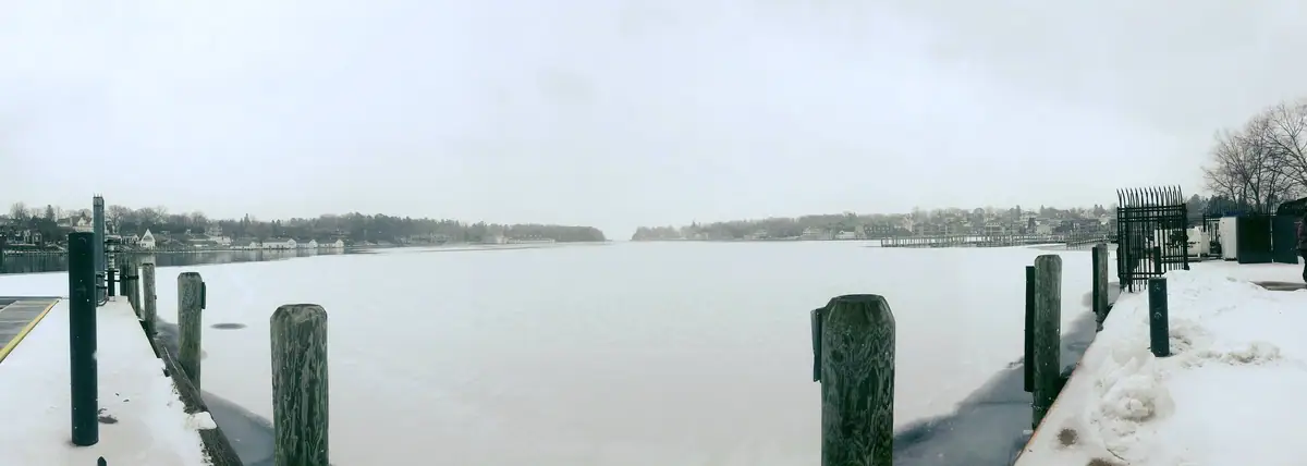 A snow-covered dock frames a broad frozen body of water with houses and trees along the far shore.