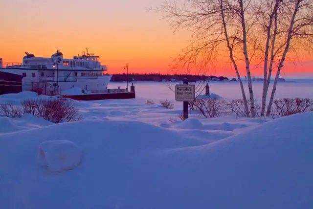 A white ferry beside a snowy waterfront at sunrise, with a park sign among leafless trees.