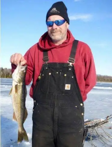 A man in winter clothing holds a large fish beside a frozen body of water.