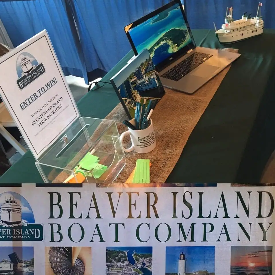 Promotional booth display with a model passenger vessel, laptop, raffle box, sign, pens, and brochures.