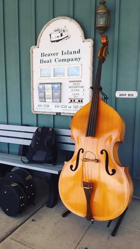 An upright bass and instrument cases sit beneath a ferry-company sign beside a bench and drop box.