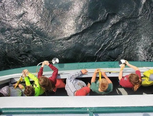 Children lean over a green railing on a boat deck and look down at the rippling water.