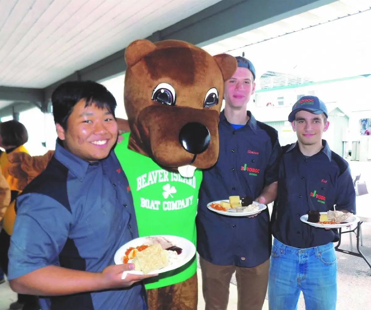 Three young men and a brown beaver mascot hold plates of food beneath a covered structure.