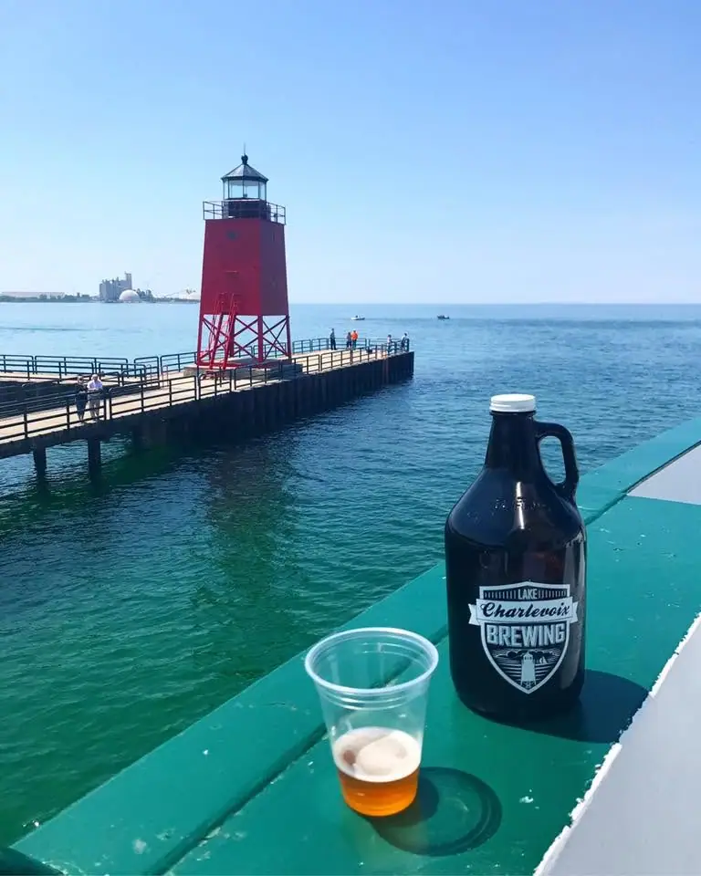 A red lighthouse stands on a pier beyond a beer growler and plastic cup on a turquoise deck.