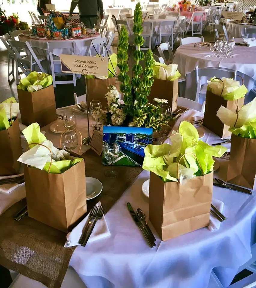A decorated banquet table holds gift bags, place settings, flowers, and a small boat photograph.