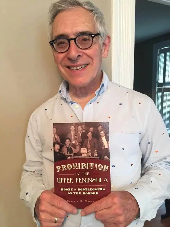 Smiling gray-haired man in glasses holds a book with a dark red cover.