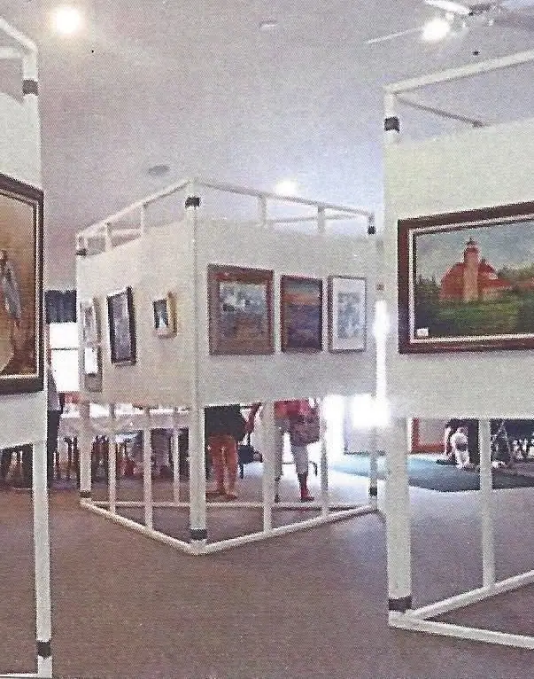 Interior exhibition space with framed artworks displayed on white partition walls and several visitors.