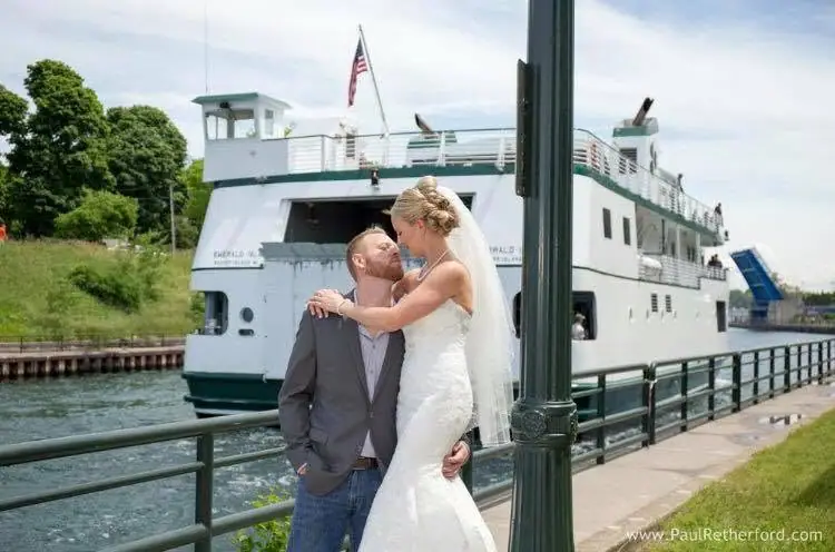 A bride and groom pose beside a large white ferry at a waterfront railing.
